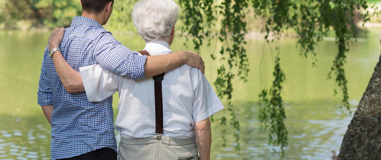 Ein älterer Herr und sein Pfleger stehen Arm in Arm an einem See.