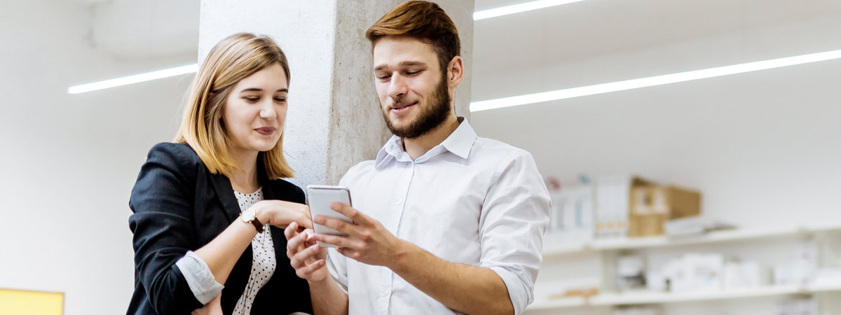 Eine junge Frau und ein junger Mann schauen gemeinsam und gut gelaunt auf ein Smartphone, welches der Mann in der Hand hält.