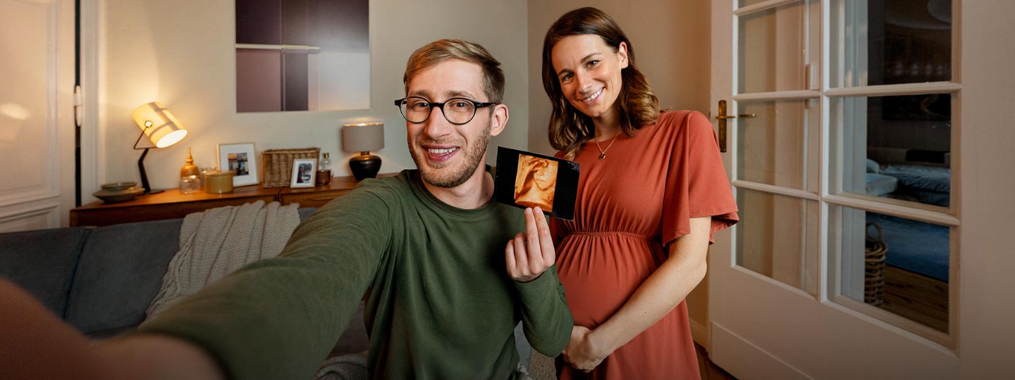 Ein Mann und eine Frau lächeln zusammen in die Kamera. Die Frau ist schwanger, der Mann hält das Ultraschallbild hoch.