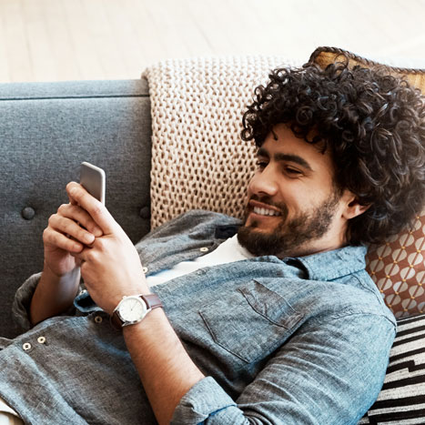 Ein junger Mann liegt auf einem Sofa und schaut lächelnd auf sein Smartphone.