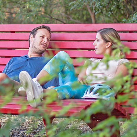 Ein Mann und eine Frau sitzen auf einer roten Bank in der Natur und unterhalten sich.