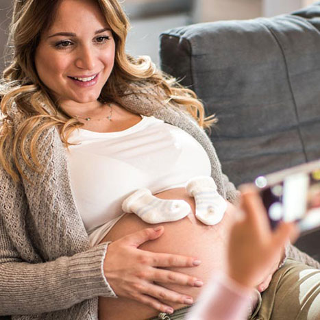 Eine Schwangere Frau sitz auf einer Couch und wird von einer Freundin mit dem Handy fotografiert.