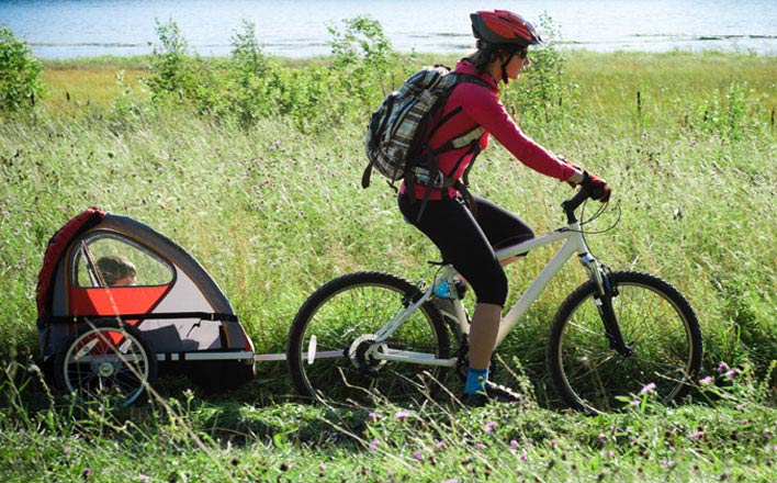 Eine Frau fährt mit ihrem Fahrrad und Kind im Anhänger in einer sonnigen Seelandschaft.