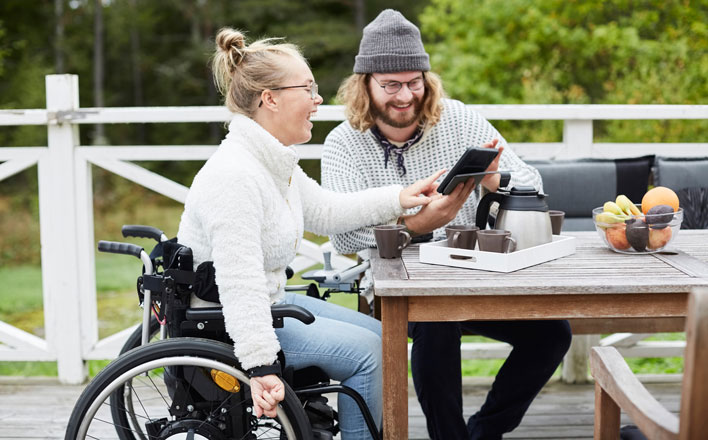Eine Frau im Rollstuhl und ihr Pfleger schauen gemeinsam auf ein Tablet und freuen sich.