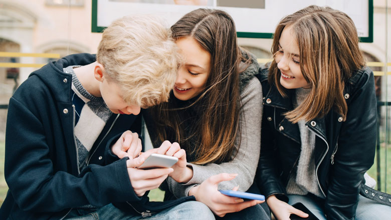 Ein Junge und zwei Mädchen sitzen zusammen und schauen auf ein Smartphone.