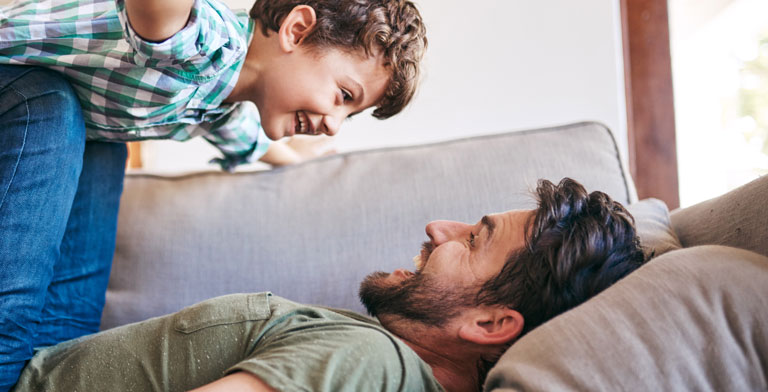 Gut versichert bei der KNAPPSCHAFT. Ein Vater spielt zu Hause auf dem Sofa mit seinem Sohn.