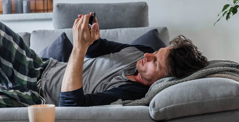 App für Long Covid – FIMO. Ein junger Mann liegt krank auf seinem Sofa und benutzt die FIMO Long Covid App