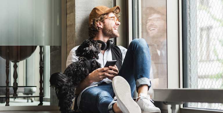 Newsletter der KNAPPSCHAFT. Ein junger Mann sitzt mit seinem Hund an einem Fenster und freut sich über den Newsletter der KNAPPSCHAFT.