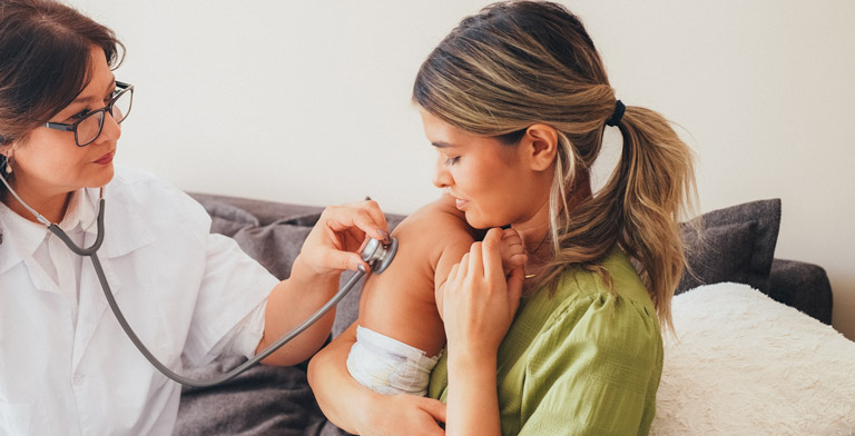 Die KNAPPSCHAFT und Ihre Hebamme. Die Hebamme untersucht das Baby auf dem Arm der Mutter mit einem Stethoskop.