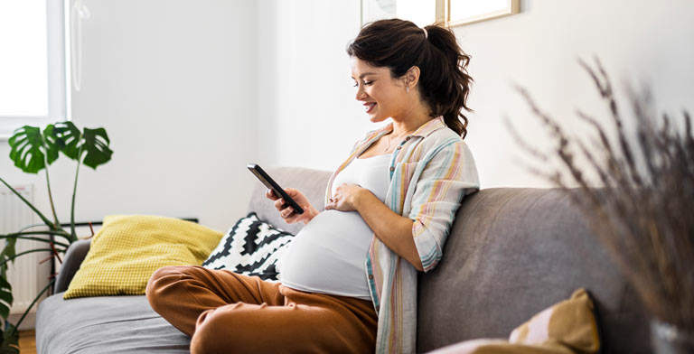 Gesund von Anfang an. Eine Schwangere sitzt auf dem Sofa und nutzt auf ihrem Smartpohne die App Lebensstark ernährt.