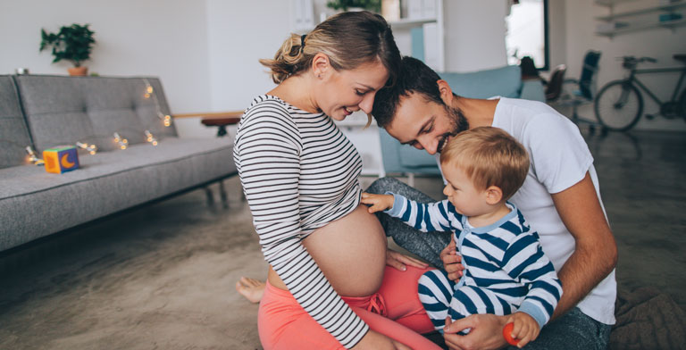 Mutterschaftsgeld. Eine Familie im Wohnzimmer. Das Kind zeigt auf den Bauch der schwangeren Mutter - diese erhält Mutterschaftsgeld.