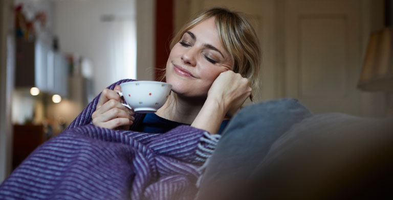 Gesund werden. Eine Frau liegt eingehüllt in eine Decke, mit einer Tasse Tee in der Hand, auf einem Sofa - sie möchte gesund werden.