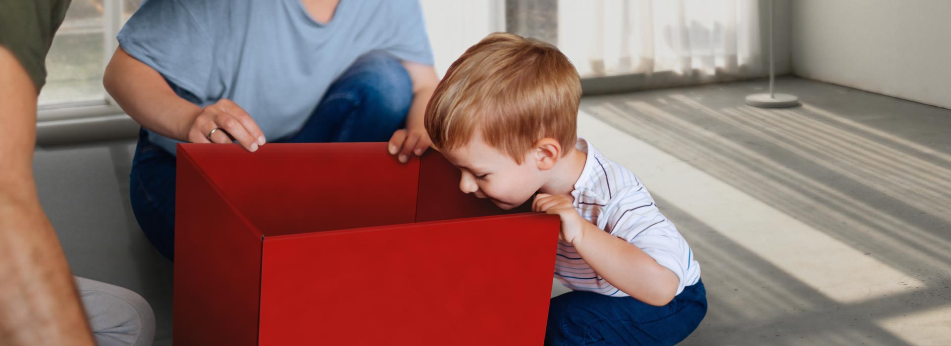 Was steckt in Ihrem Gesundheitspaket? Ein kleines Kind schaut, zusammen mit seinen Eltern, neugierig in eine große rote Paketbox.