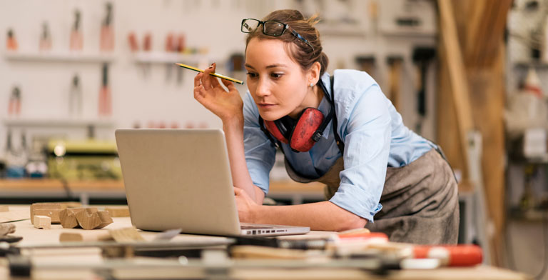 Krankenversicherung für Arbeitnehmer. Eine Tischlerin schaut in der Werkstatt konzentriert auf einen Laptop und informiert sich über die Krankenversicherung für Arbeitnehmer.