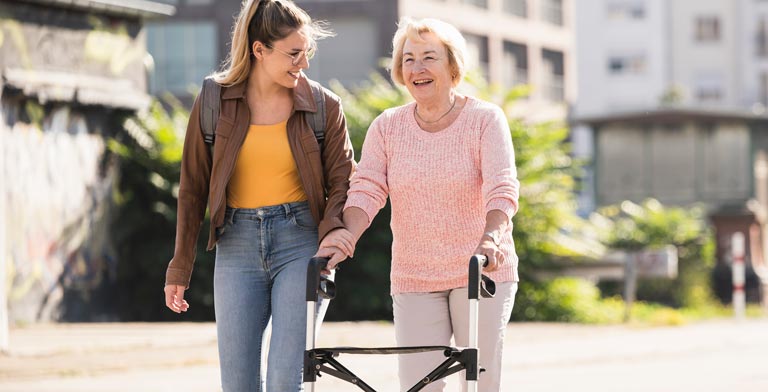 Umfrage zu Hilfsmitteln. Eine Seniorin mit einem Rollator und eine junge Frau bei einem Spaziergang.
