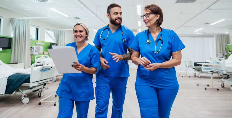 Die sind für Sie da. Drei lächelnde Pfleger in blauer Kleidung laufen nebeneinander in einem Krankenhaus der KNAPPSCHAFT.
