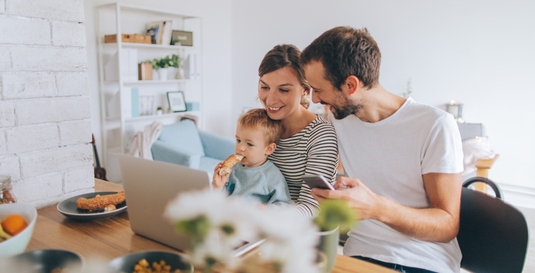 Familienversicherung. Ein junges Paar sitzt mit ihrem Sohn auf dem Schoß am Frühstückstisch und informiert sich am Handy und am Laptop zur Familienversicherung.