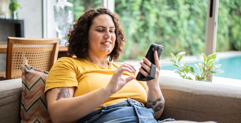 Freiwillige Krankenversicherung. Frau sitzt lächelnd auf dem Sofa und informiert sich auf ihrem Smartphone über die freiwillige Mitgliedschaft der KNAPPSCHAFT.