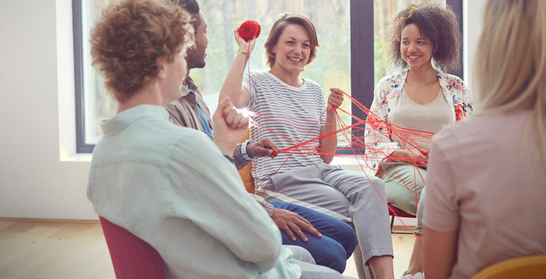 Selbsthilfeförderung. Gruppe von Menschen sitzt zur Selbsthilfeförderung in einem Kreis. Sie werfen sich lächelnd ein rotes Knäul Wolle zu.