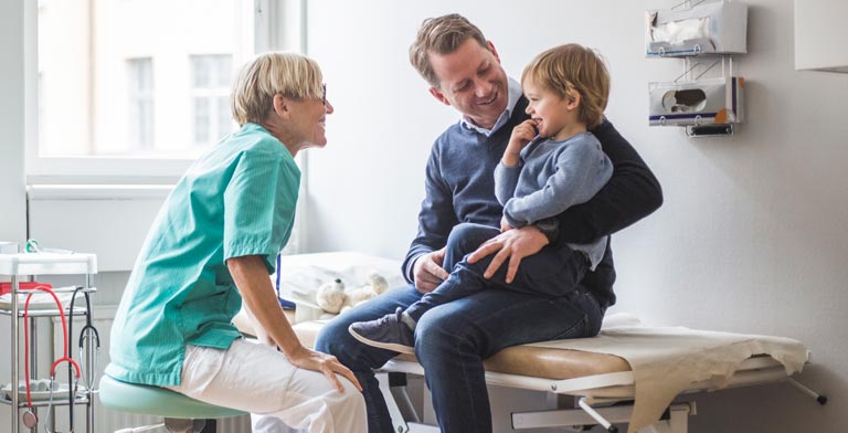 Im Krankenhaus. Ein Vater sitzt mit Kind auf dem Schoß auf der Behandlungsliege aufgrund einer stationären Behandlung. Die Ärztin sitzt gegenüber und lächelt.