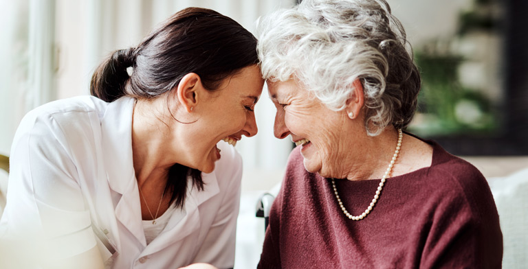 Stationäre Pflege. Eine Seniorin und eine Pflegerin sitzen nebeneinander und lachen Stirn an Stirn.
