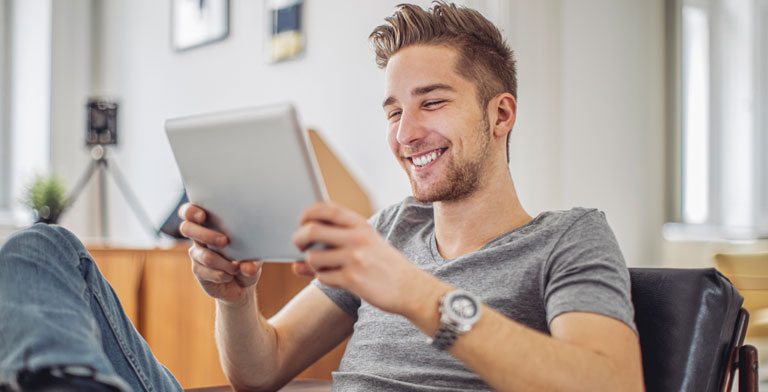 Alles rund um die Online-Psychotherapie. Ein junger Mann sitzt mit einem Tablet auf dem Sofa und nimmt die Online-Psychotherapie in Anspruch.