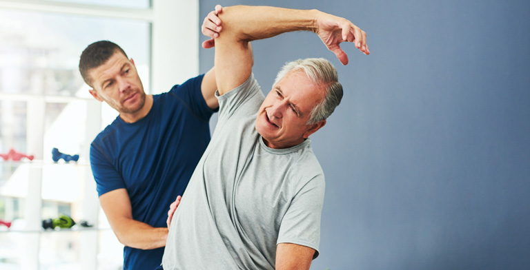 Physiotherapie. Ein Mann sitzt bei der Physiotherapie. Ein weiterer Mann steht hinter ihm und hält seine Schulter fest, währenddessen er ihn untersucht.