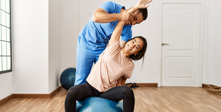 Therapien und besondere Behandlungsfälle. Ein junge Frau bei ihrer Therapie in Form einer Physiotherapie. Sie sitz auf dem Boden, währenddessen der Mann hinter ihr einen ihrer Arme über ihren Kopf auf die andere Seite führt.