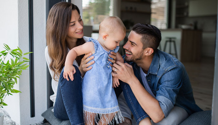 AktivBonus junge Familie Ein Mann und eine Frau lächeln ein Kind an und halten es in den Armen. Sie sitzen dabei nebeneinander auf dem Boden.