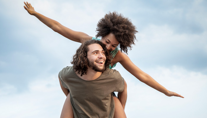 AktivBonus und SportBonus Eine junge Frau macht eine Fliegerpose auf dem Rücken ihres Freundes. Sie freuen sich über den Aktivbonus.