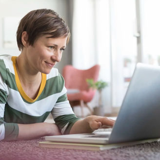 Eine lächelnde Frau liegt zuhause auf dem Boden vor einem aufgeklappten Laptop Eine lächelnde Frau liegt zuhause auf dem Boden vor einem aufgeklappten Laptop