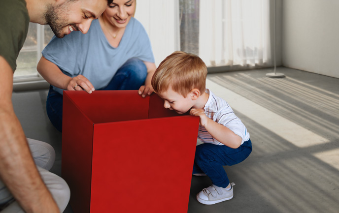 Gesundheitspaket-Test Ein kleines Kind schaut, zusammen mit seinen Eltern, neugierig in eine große rote Paketbox.