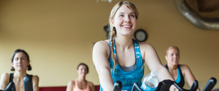 Lisa (24) AktivBonus Ein junge Frau trainiert auf einem Ergometer Fahrrad in einem Fitnessstudio und lächelt fröhlich.